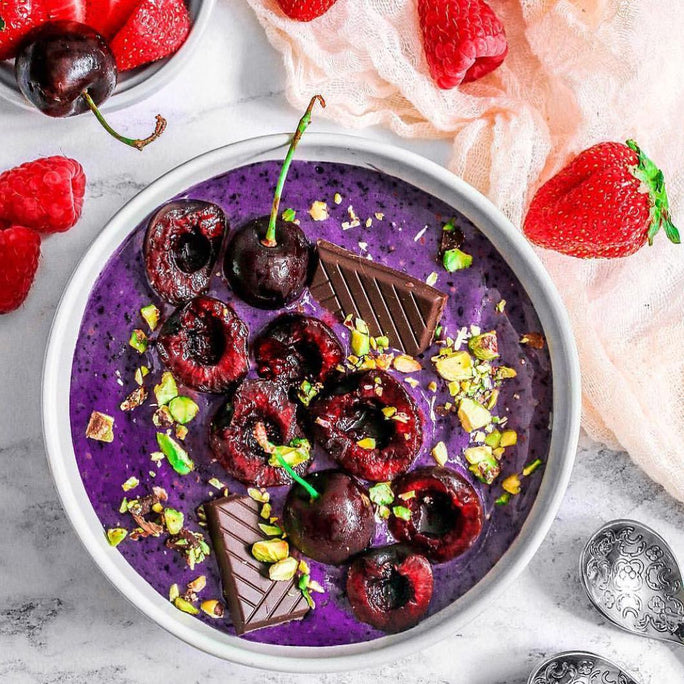 A bright purple smoothie bowl topped with deep red cherries and dark chocolate