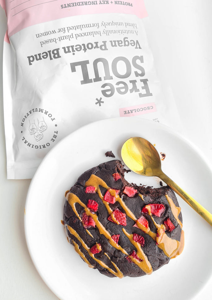 a protein chocolate cookie with a bite taken out of it, drizzled in almond butter and strawberries next to a bag of free soul vegan protein blend 