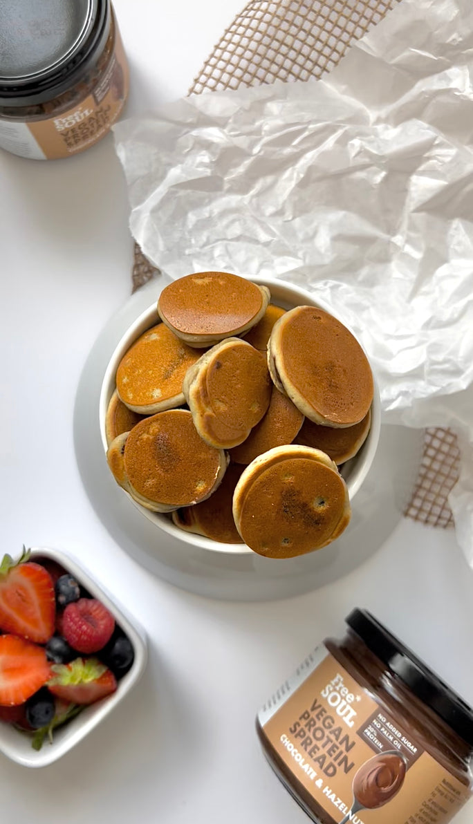 a bowl of mini pancakes with two jars of free soul vegan protein chocolate spread and a bowl of strawberries and blueberries