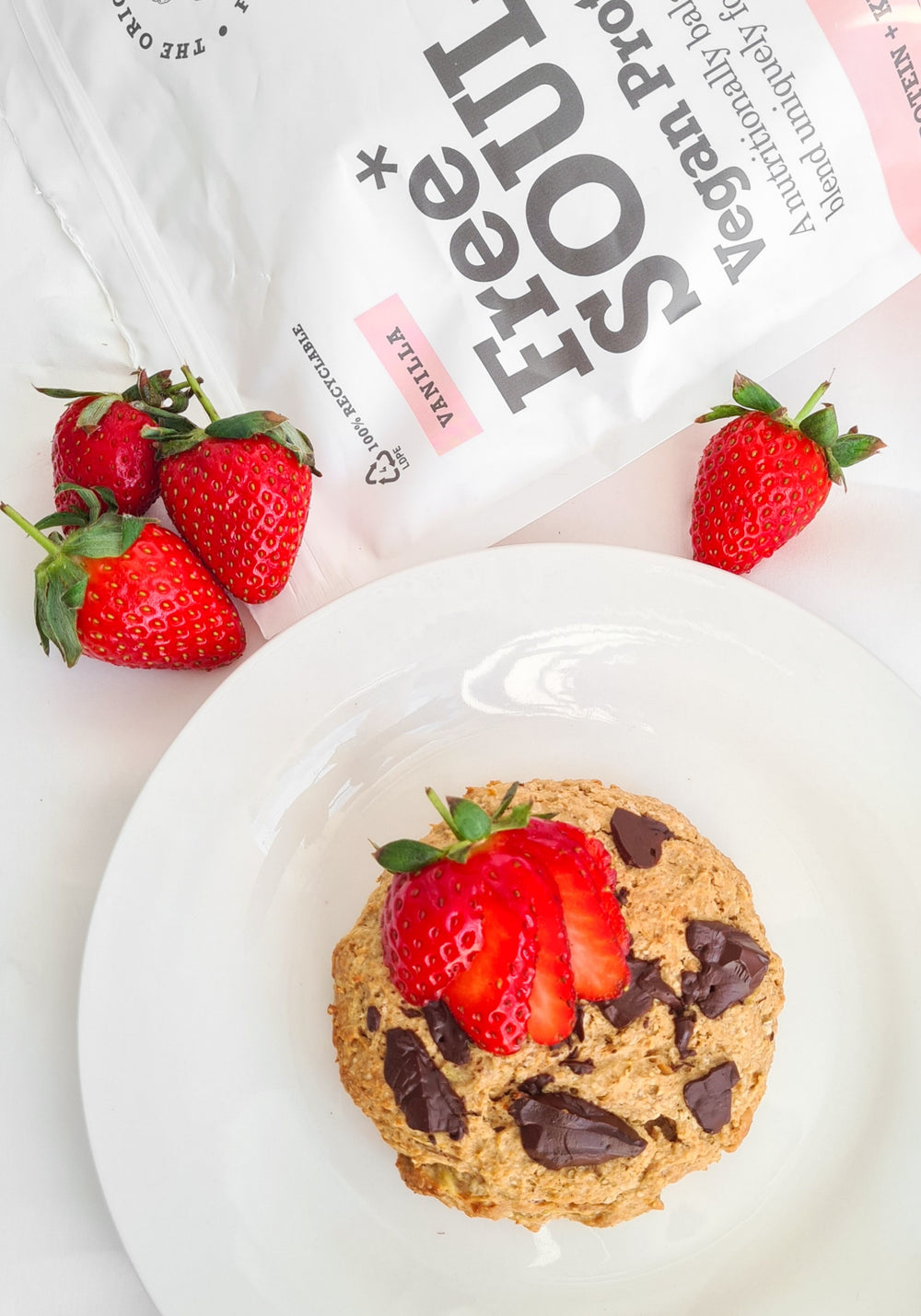 Chocolate chip cookie with strawberries on top on a white plate next to Free Soul packaging and strawberries