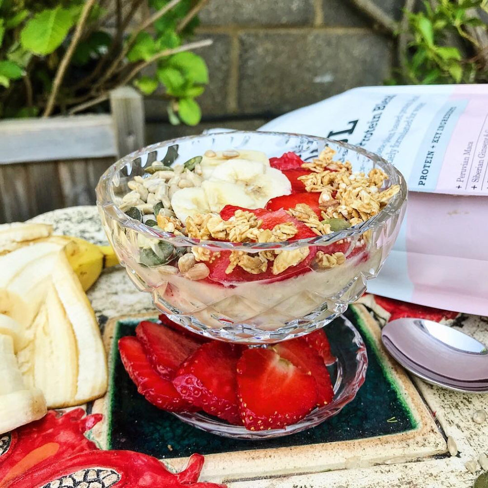A glass bowl filled with yoghurt, chopped bananas, strawberries, pumpkin seeds and granola