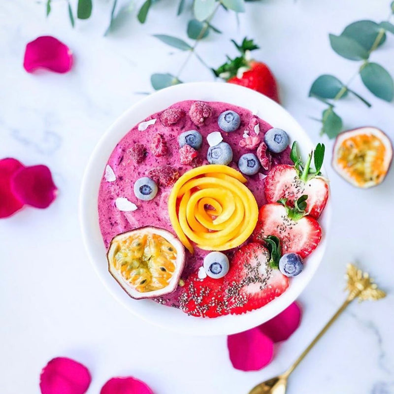 A white bowl filled with purple smoothie, fresh passionfruit and sliced mango shaped into a rose