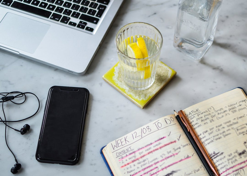 A glass of water with two slices of lemon on a table with a phone, laptop and planner